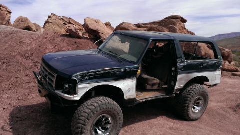 1994 Ford Bronco - This modified Ford Bronco is parked on a rocky, desert-like landscape with its driver's door open.