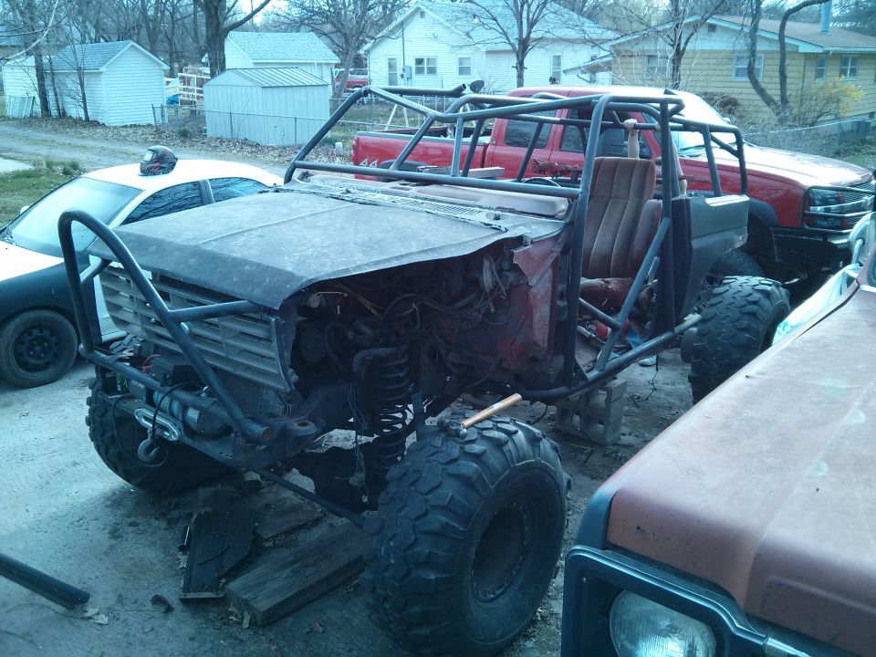 Custom off-road buggy project with roll cage, large tires, and exposed engine bay. Another red truck and a white car are in the background
