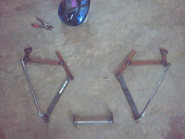 Two metal brackets and a connecting bar on a concrete floor, with a welding mask and pliers nearby