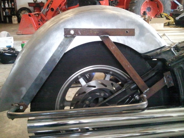 Motorcycle rear wheel with custom metal fender and exhaust pipes. The fender is unfinished metal