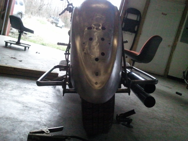 Rear view of a custom motorcycle with a raw metal fender and dual exhaust pipes