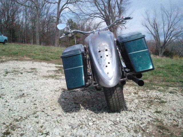 Motorcycle with two green saddlebags and a shiny metal fender. A blue truck is visible in the background