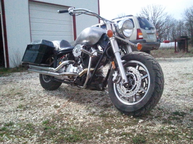 Custom motorcycle with saddlebags and chrome exhaust parked on gravel. SUV visible in background