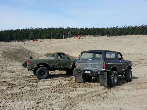 1979 Ford Bronco - Two vehicles sit on a sandy terrain, with a forest line visible in the distance under a clear sky.