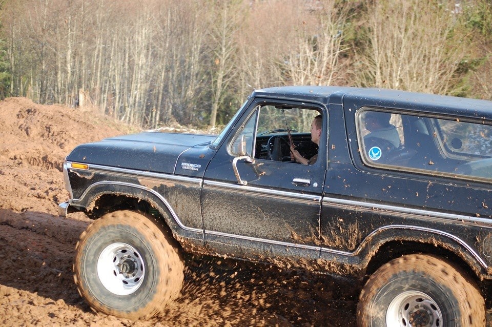 Black Ford Bronco driving through mud, tires kicking up dirt. Driver visible inside