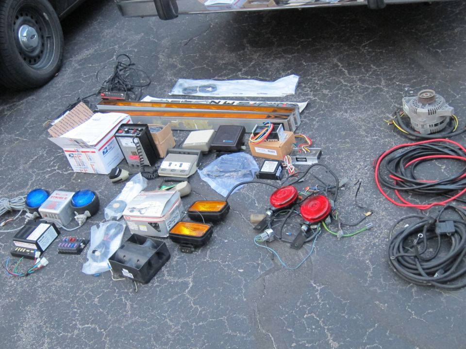 Assorted vehicle emergency lights, sirens, wiring, and an alternator laid out on asphalt