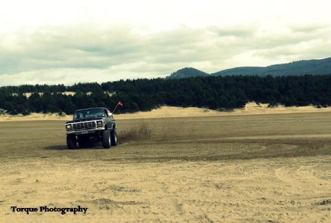 1979 Ford Bronco - A black Ford Bronco drives across a sandy terrain with a forest and hills in the distance.
