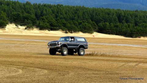 1979 Ford Bronco - A dark SUV with a lifted suspension drives across a sandy landscape with a forest in the background.
