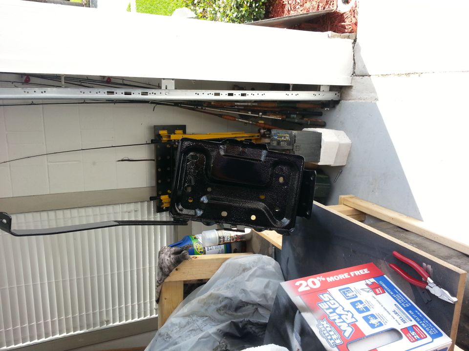 Black metal battery tray with yellow brackets, sitting on wood. Pliers and a SuperMotors ad are nearby