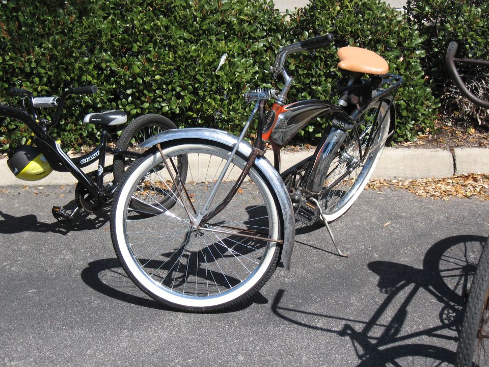 Chrome cruiser bicycle with tan seat and whitewall tires, parked next to a black bicycle with a yellow helmet