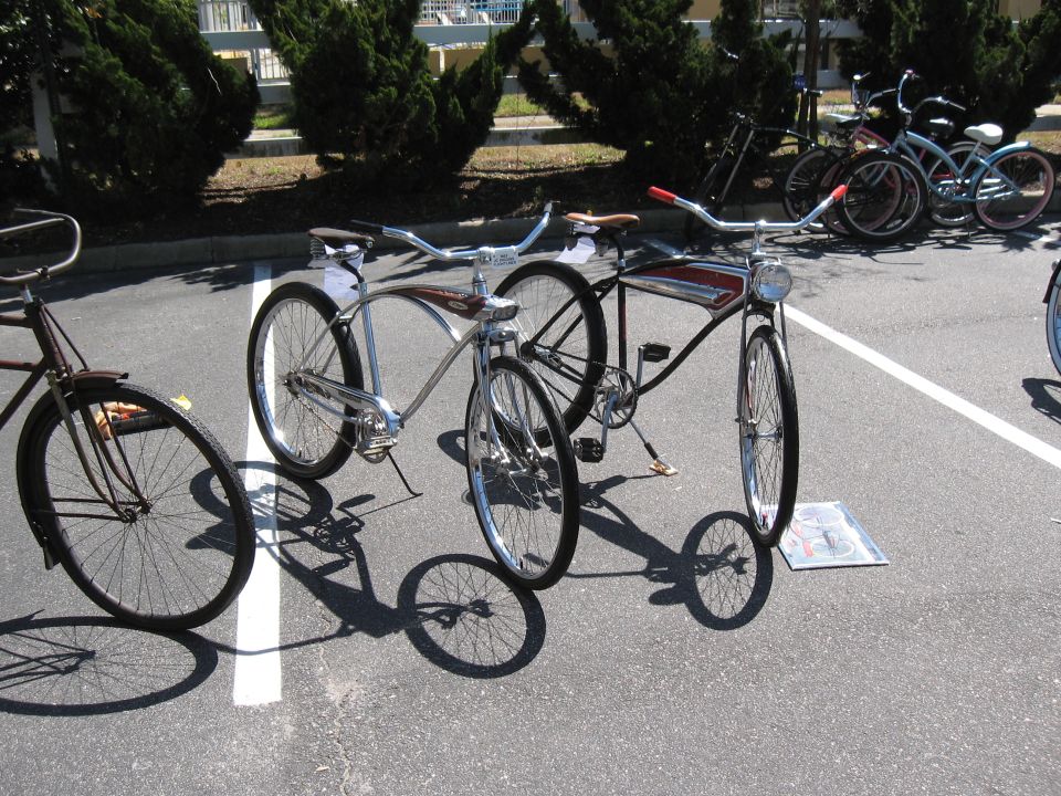 Several cruiser bicycles parked on asphalt, with trees and a fence in the background