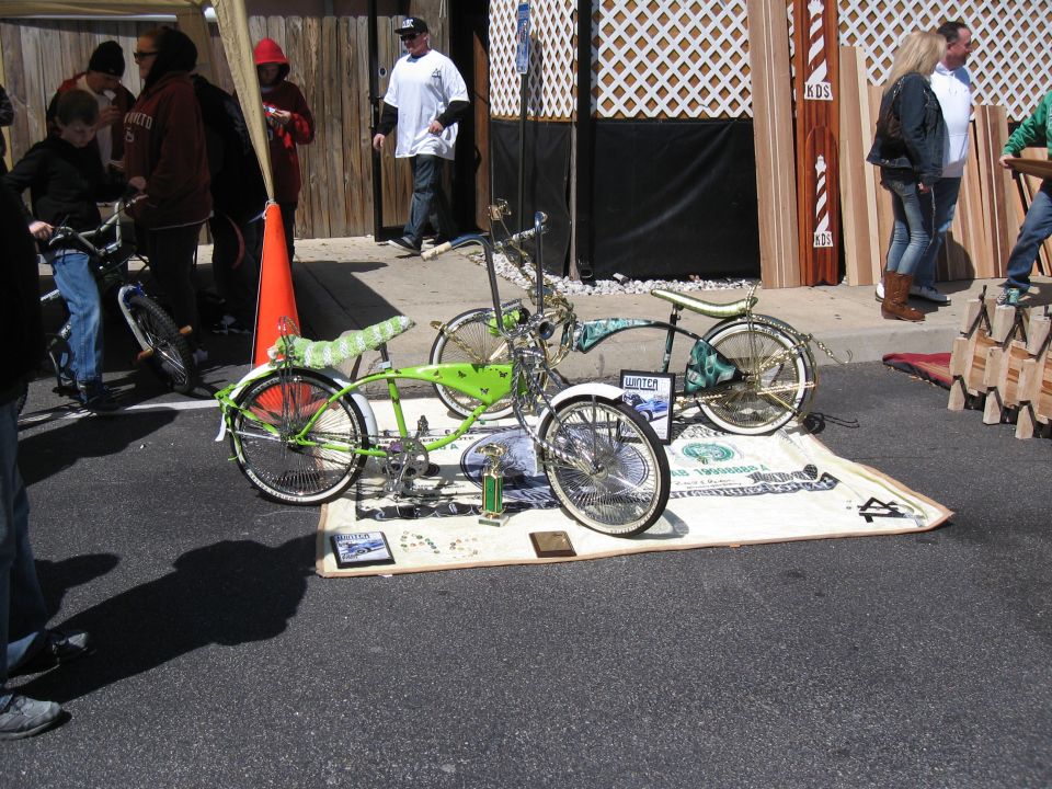 Two custom lowrider bicycles with green and gold accents displayed on a large dollar bill mat
