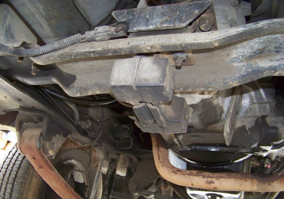 Underside view of a vehicle's frame, transmission, and exhaust pipe. Shows some rust and wear