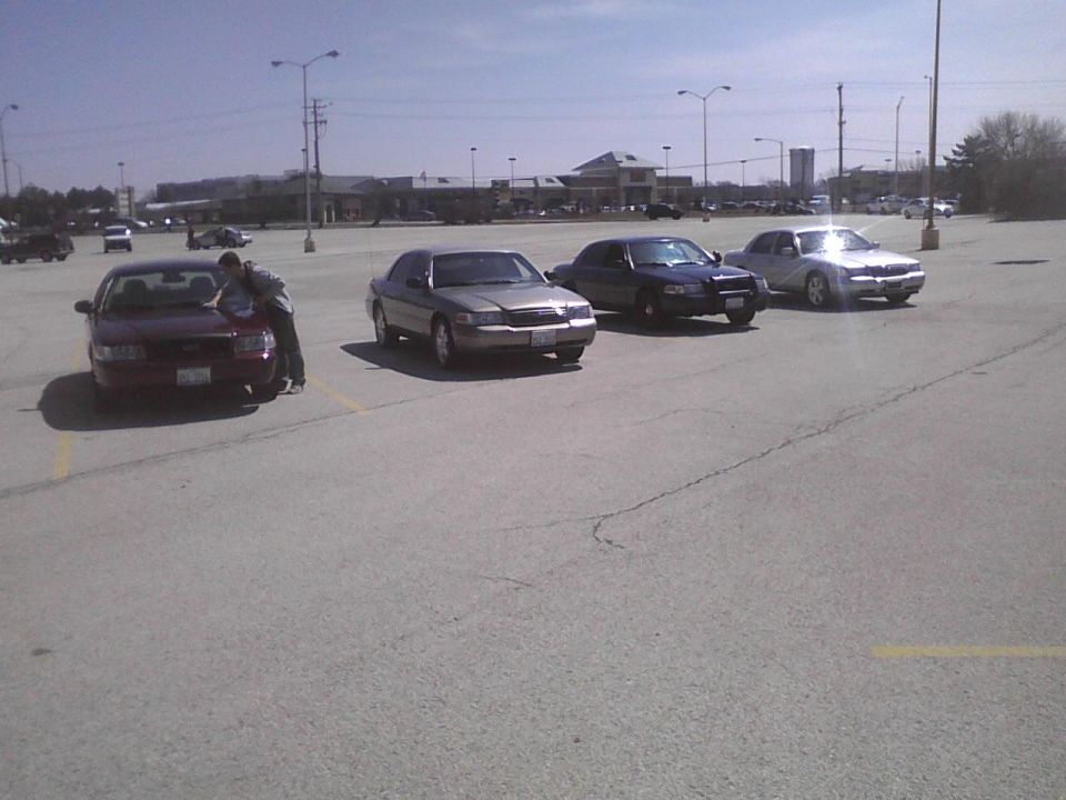 Four sedans parked in a lot, one is a maroon 2003 Ford Crown Victoria with a person leaning on it