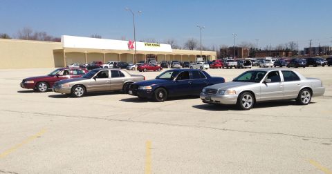2005 Ford Crown Victoria - Several Ford Crown Victorias, some appearing to be police cruisers, are parked in a lot outside a store.