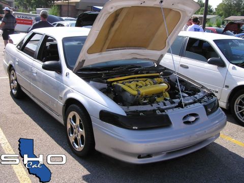 1993 Ford Taurus - A silver 1993 Ford Taurus is displayed with its hood open, a vibrant yellow engine at a car show.