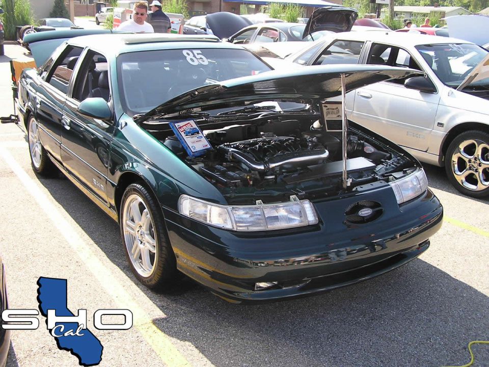 Green Ford Taurus with hood open, showing engine. Another car is parked to the right. Number 85 is on the windshield