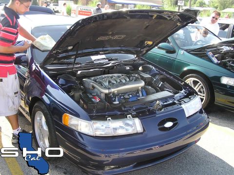 1993 Ford Taurus - A man cleans the windshield of a blue Ford Taurus SHO with its hood open, revealing the engine.