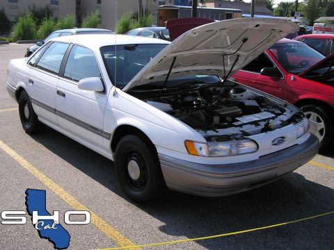 1993 Ford Taurus - A white sedan is parked with its hood open, alongside another vehicle in a parking lot.