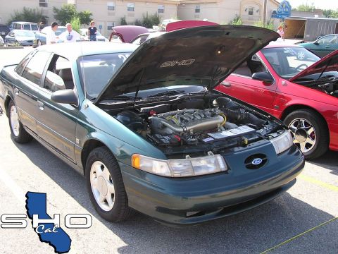 1993 Ford Taurus - A green 1993 Ford Taurus SHO is displayed with its hood open, revealing the V6 engine, at a car show with other vehicles present.