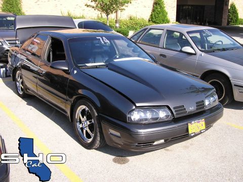 1993 Ford Taurus - A dark sedan with a prominent hood scoop and aftermarket wheels is parked outdoors, with another vehicle visible behind it.