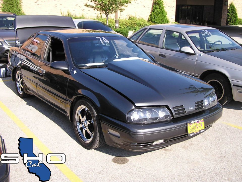 Dark sedan with a hood scoop and custom wheels parked next to a silver car. A yellow license plate reads 'SHONEM'