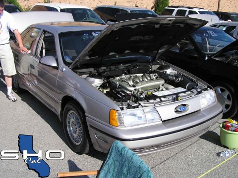 1993 Ford Taurus - A silver sedan with its hood open, revealing the engine, is parked outdoors with other vehicles.