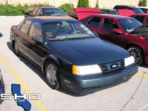 1993 Ford Taurus - A dark green sedan is parked in a lot with other vehicles, some with hoods open.