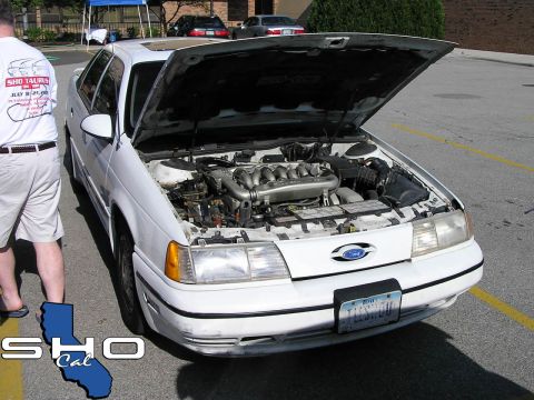 1993 Ford Taurus - A white 1993 Ford Taurus is parked with its hood open, revealing the V6 engine. The car is viewed from the front.