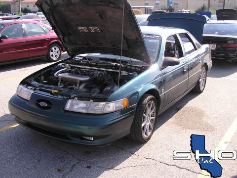 1993 Ford Taurus - A green Ford Taurus SHO is parked with its hood open, revealing the engine bay. Another vehicle is visible to the left.