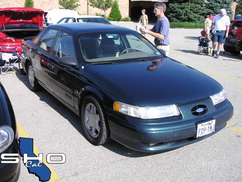 1993 Ford Taurus - A dark green Ford Taurus is parked in a lot with other vehicles and people around.