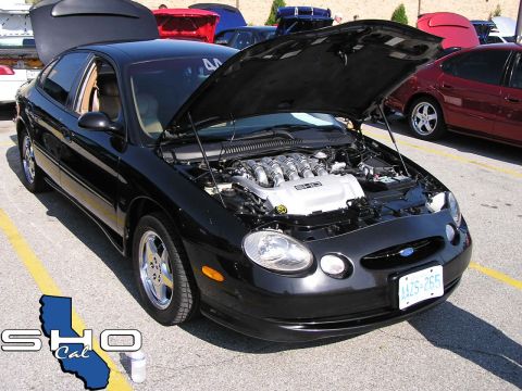 1993 Ford Taurus - A black Ford Taurus is displayed with its hood open, revealing the engine compartment. Other vehicles are parked nearby.