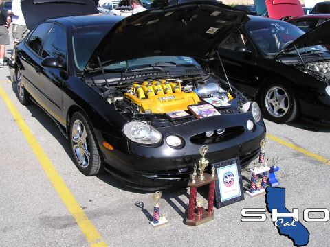 1993 Ford Taurus - A black Ford Taurus with its hood open, displaying a custom yellow V8 engine and trophies, parked at an outdoor event.