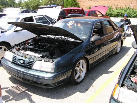 1993 Ford Taurus - A dark green sedan with its hood open is parked at an outdoor event with other vehicles.