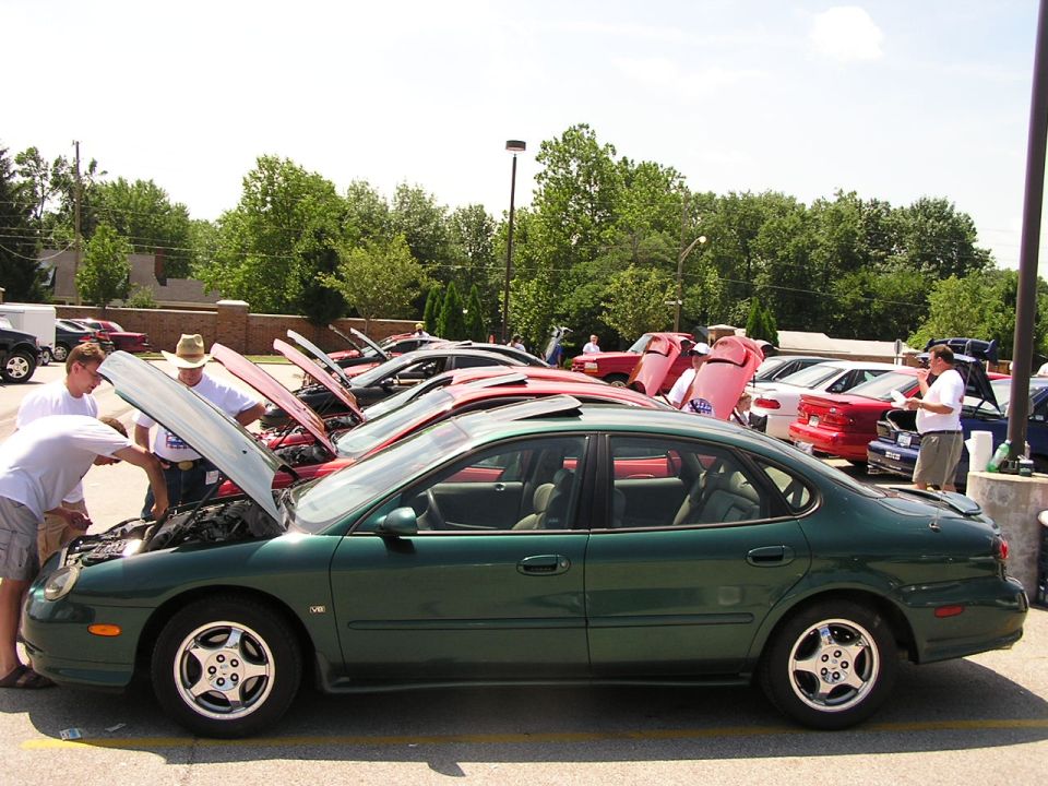 Green sedan with sunroof, side view. Other cars with hoods open in background