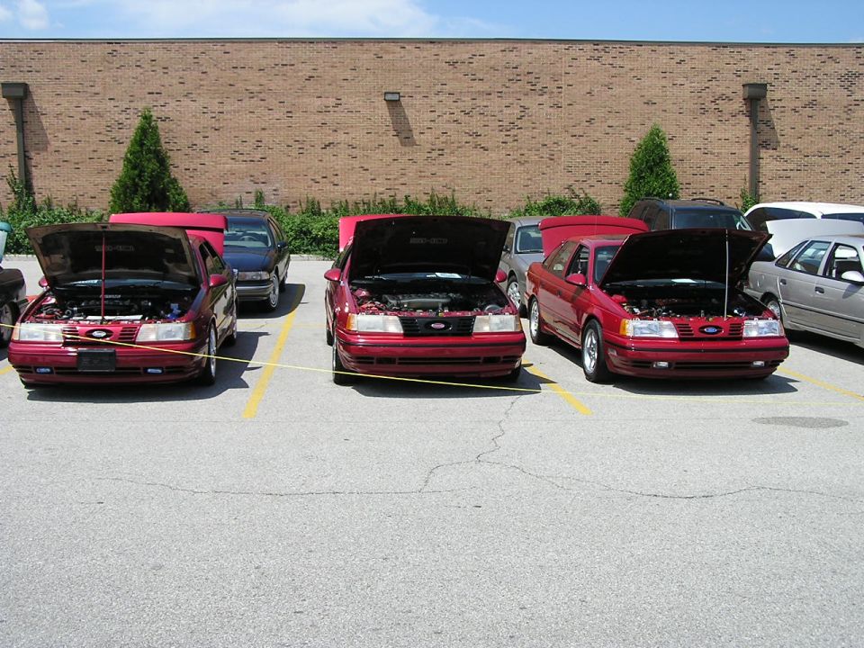 Three red Ford Taurus cars parked in a lot with hoods up. Another car is visible to the right