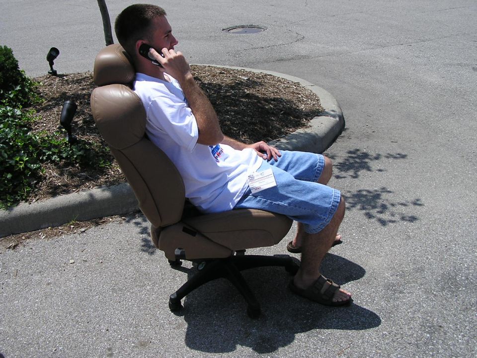 Man in white t-shirt and denim shorts sits in a tan office chair outdoors, talking on a cell phone