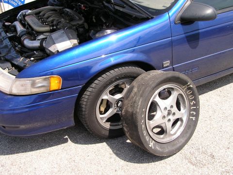 1993 Ford Taurus - A blue 1993 Ford Taurus is shown with its hood open, revealing the engine and a Hoosier drag radial tire placed beside the front wheel.