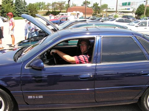1993 Ford Taurus - A young man sits in the driver's seat of a blue sedan, with other vehicles and people visible in the background.