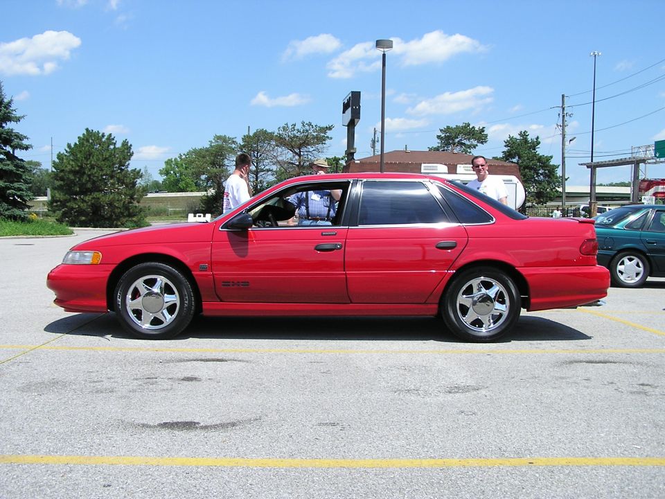 Red 1993 Ford Taurus sedan parked in lot, side view. Another car is visible to the right