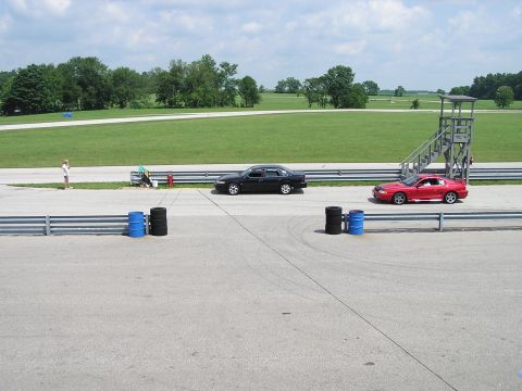 1993 Ford Taurus - Two cars are positioned on a racetrack, with a person standing nearby and a race control tower in the background.