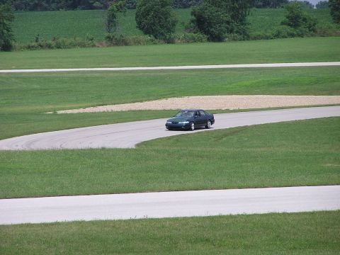 1993 Ford Taurus - A dark green 1993 Ford Taurus navigates a turn on a race track surrounded by grassy fields.