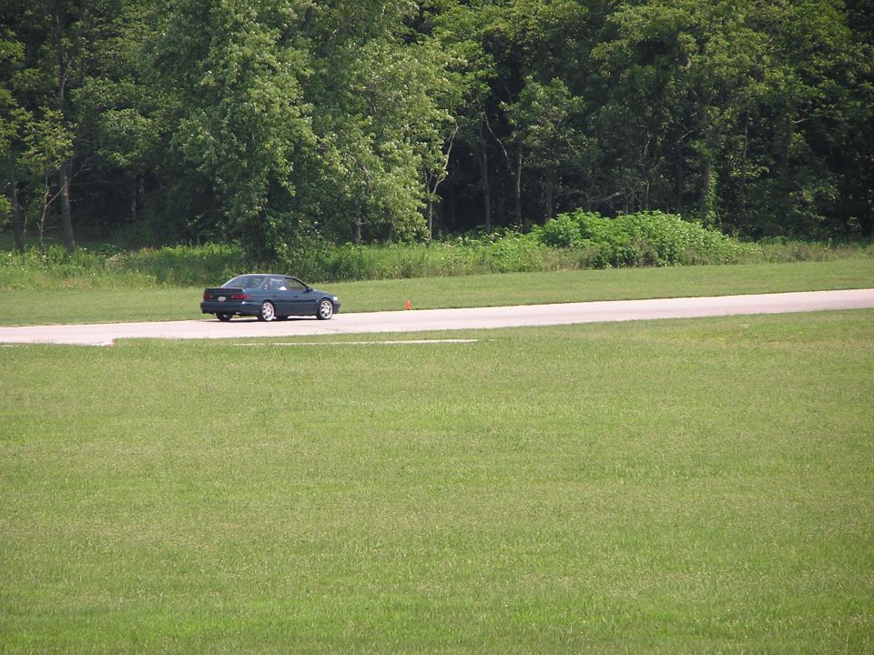 Dark sedan driving on a paved track with grassy fields and trees in the background