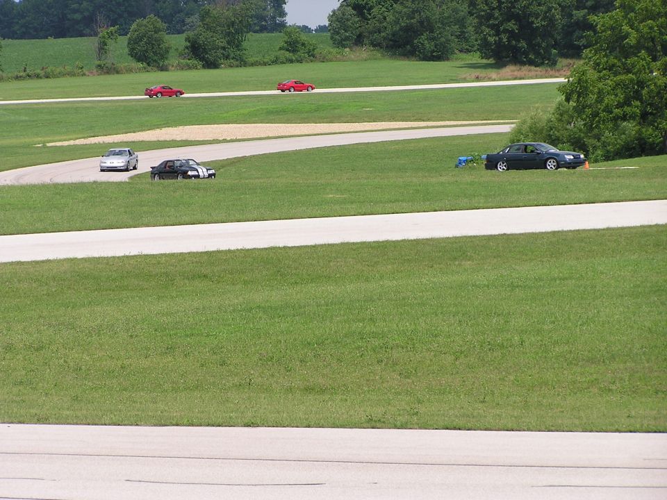 Several cars on a grassy race track, including red sports cars and a black sedan