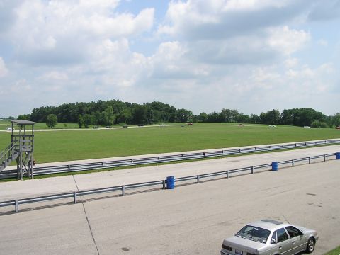 1993 Ford Taurus - A silver 1993 Ford Taurus is parked on the edge of a race track, with several other vehicles visible on the course in the background.