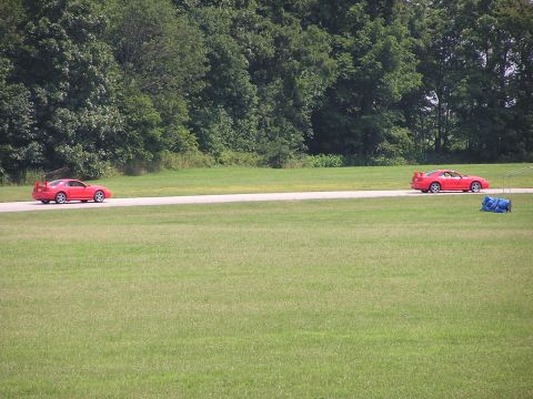 1993 Ford Taurus - Two red sports cars drive on a road surrounded by grass and trees.