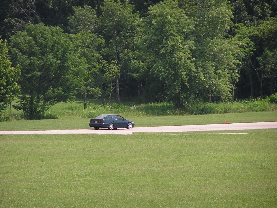 Dark sedan driving on a paved road with a grassy field and trees in the background