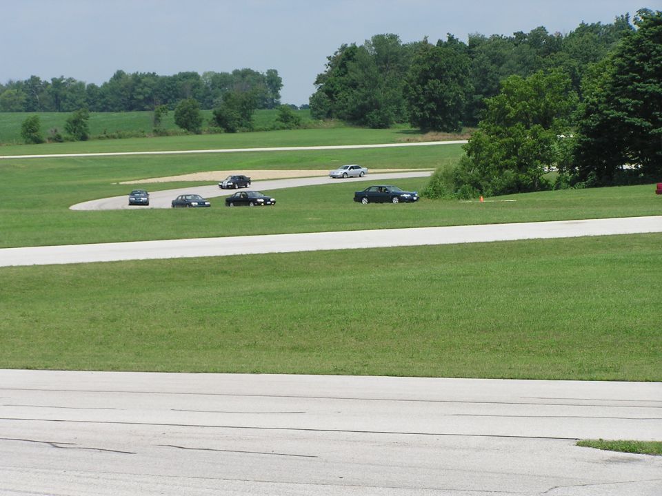 Several cars drive on a paved racetrack surrounded by grass and trees