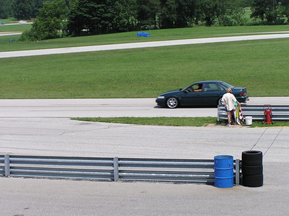 Dark green sedan driving on a racetrack with a person standing nearby holding flags