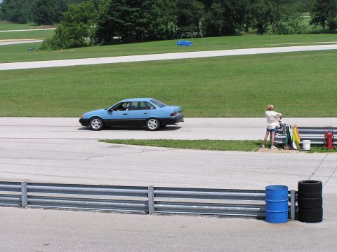 1993 Ford Taurus - A two-tone sedan drives on a track with a person attending to equipment nearby.